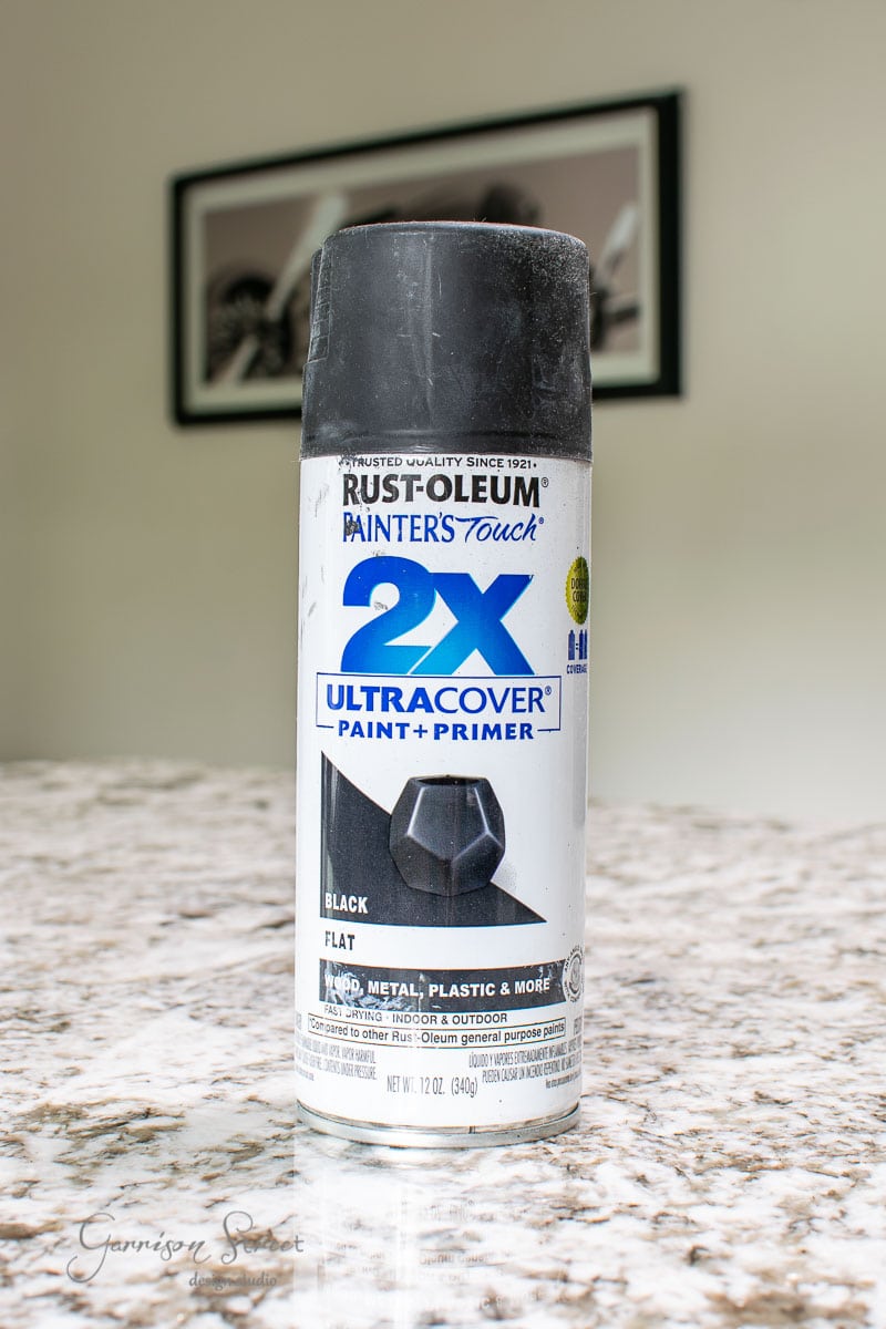 A can of Rust-Oleum Painter's Touch 2X Ultra Cover paint and primer in Luftmassa black flat is sitting on a marble countertop. The label indicates it's suitable for wood, metal, plastic, and more. A framed photo is blurred in the background.