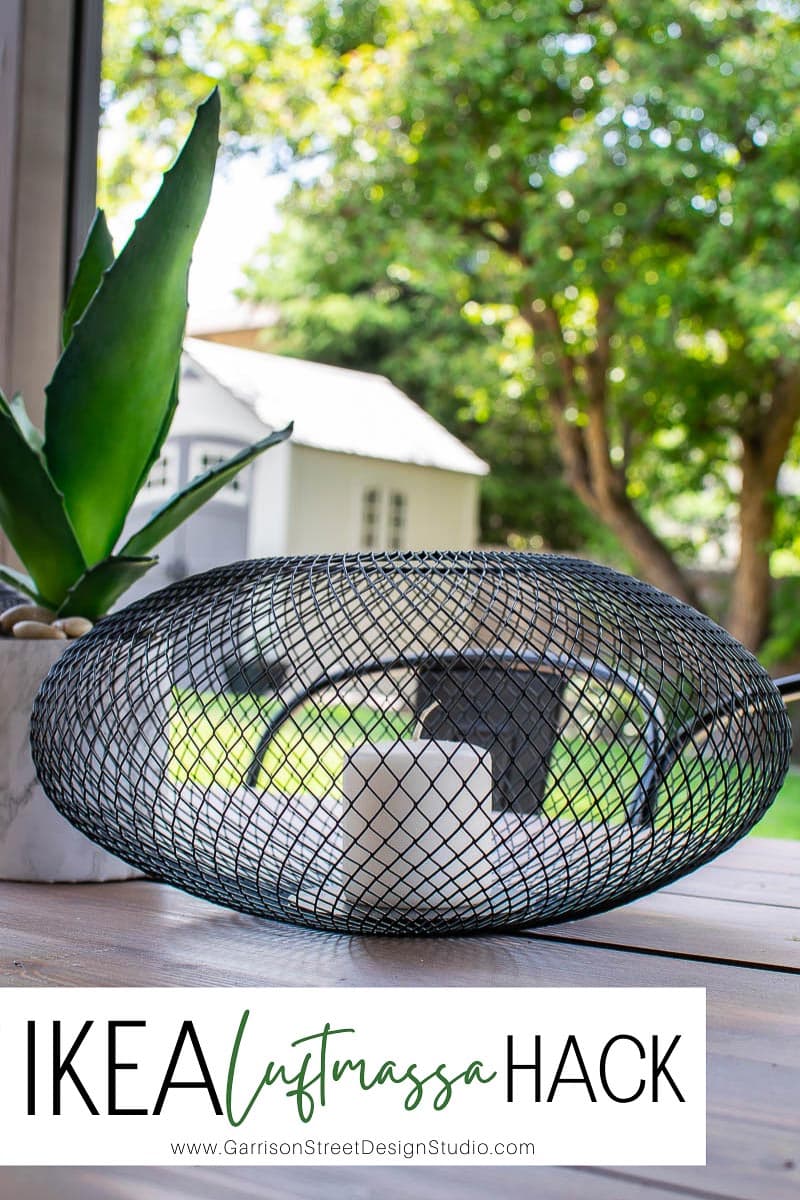 A black wire mesh bowl containing white candles sits on a table beside a vibrant green plant. The scene unfolds against a lush backdrop of greenery and a white house. Text overlay reads "IKEA Luftmassa Hack" with a website link, showcasing this stylish IKEA-inspired creation.