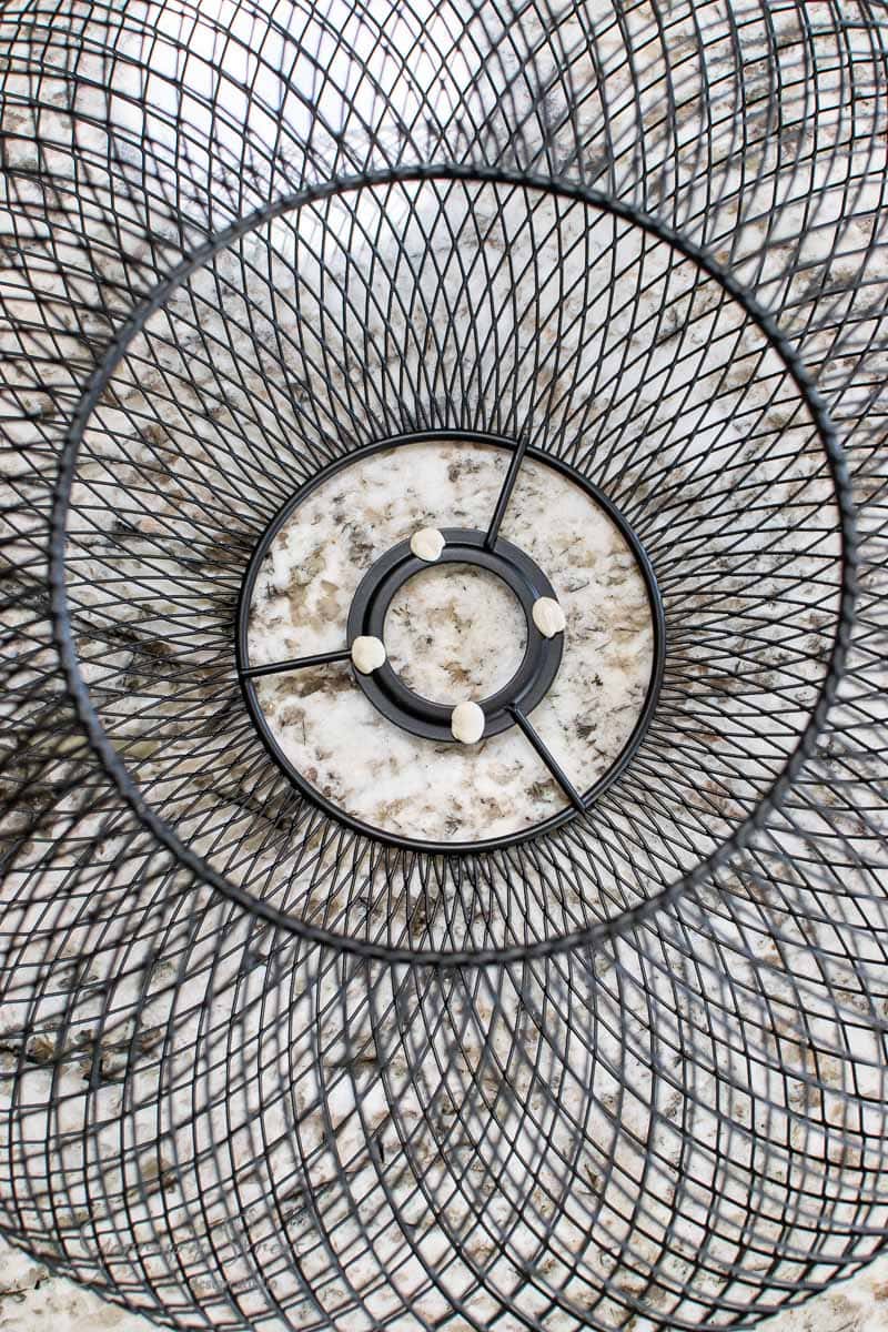Top view of a Luftmassa wire mesh trash bin on a granite surface. The IKEA bin's circular, grid-like structure creates a geometric pattern, with four small felt pads visible at the bottom. The granite surface has a speckled texture.