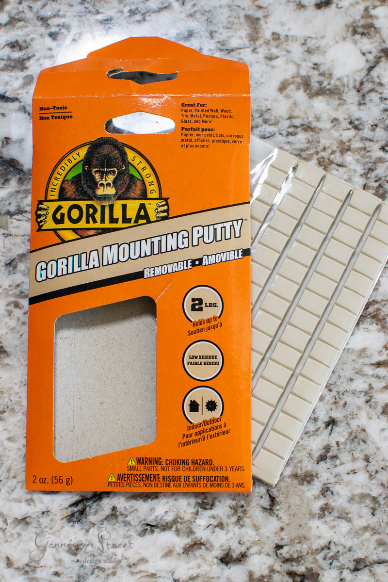 A package of Gorilla Mounting Putty rests on a stone surface. The orange box showcases the product name and attributes like being removable and non-toxic. The putty strips are arranged in a grid layout.
