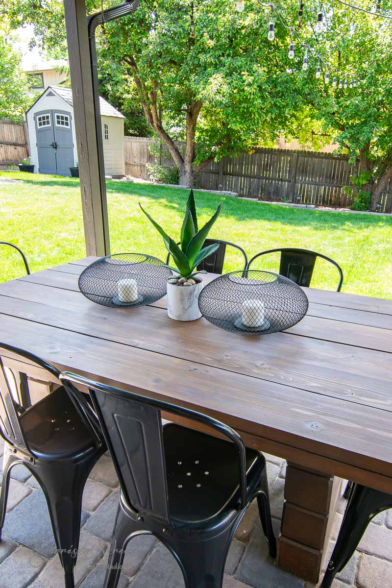 An IKEA wooden outdoor dining table with black metal chairs is set on a patio. The table features a small potted plant and two black Luftmassa wire baskets as decoration. In the background, a lush green yard and a small garden shed are visible.