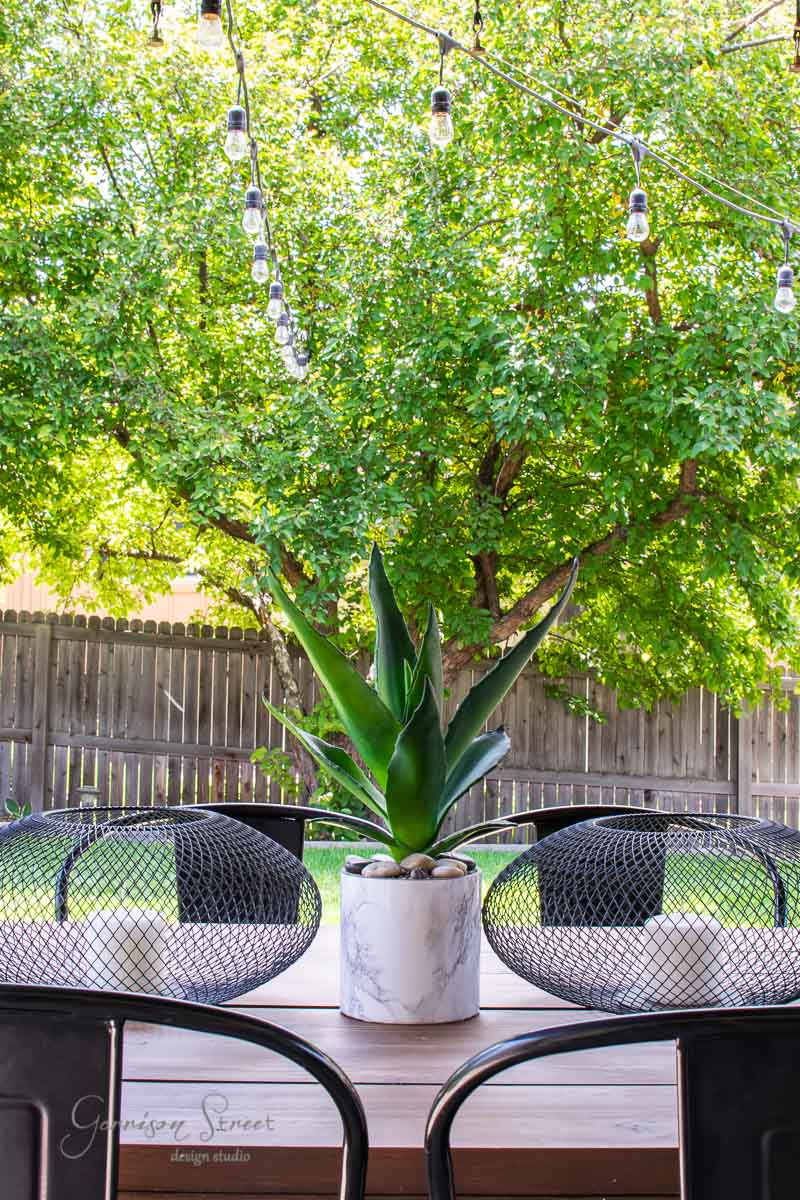 A backyard patio showcases an IKEA wooden table with a potted succulent centerpiece. Two black Luftmassa wire chairs are nestled on each side. String lights dangle overhead, while a tall, lush green tree offers a natural backdrop against the enclosing wooden fence.