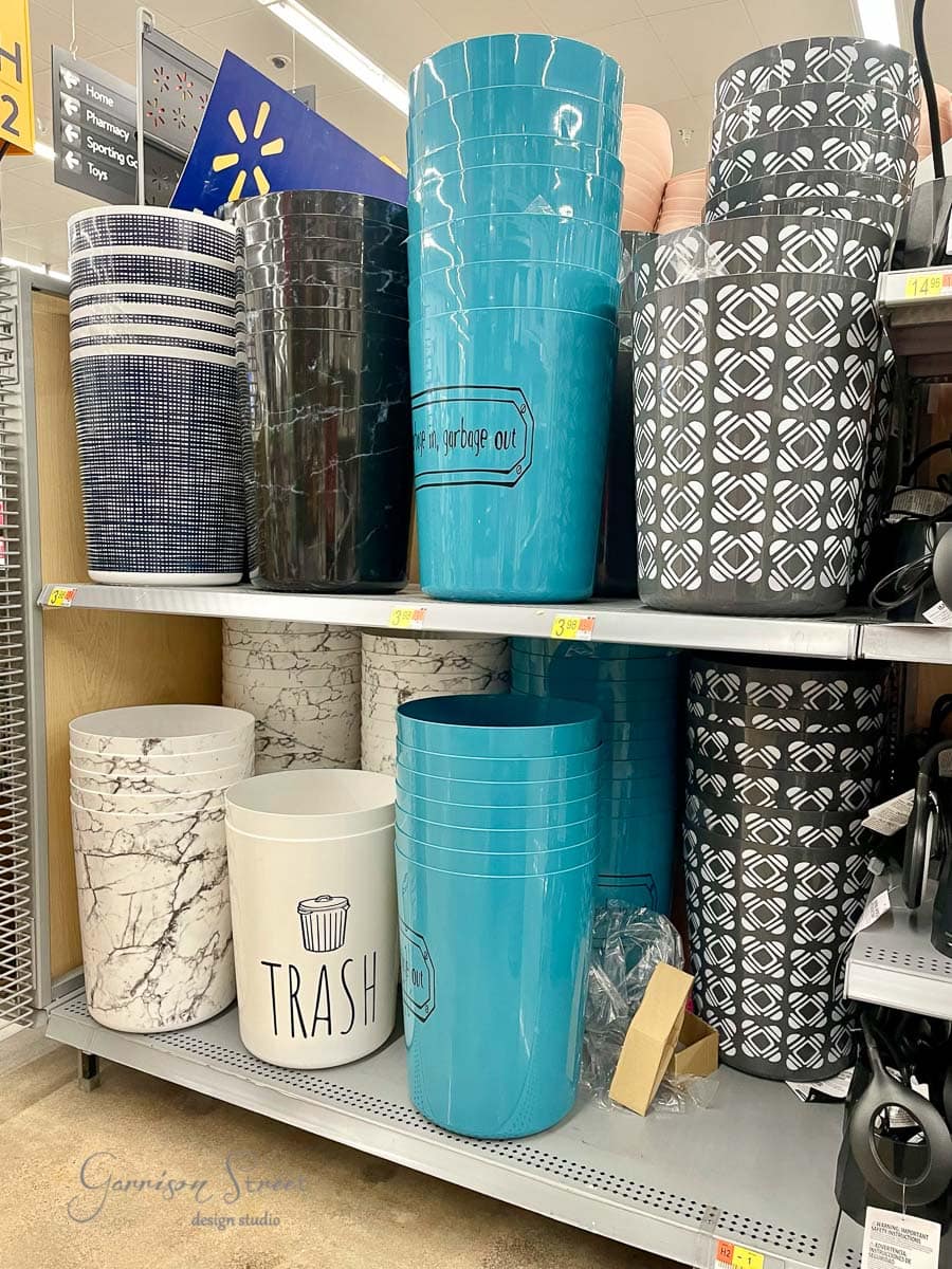 A store shelf displays various wastebaskets. The top shelf holds striped and patterned black, white, and blue wastebaskets. The bottom shelf features light blue, marble-patterned, and white bins with the word "TRASH," nestled beside a sleek concrete planter for a modern touch.