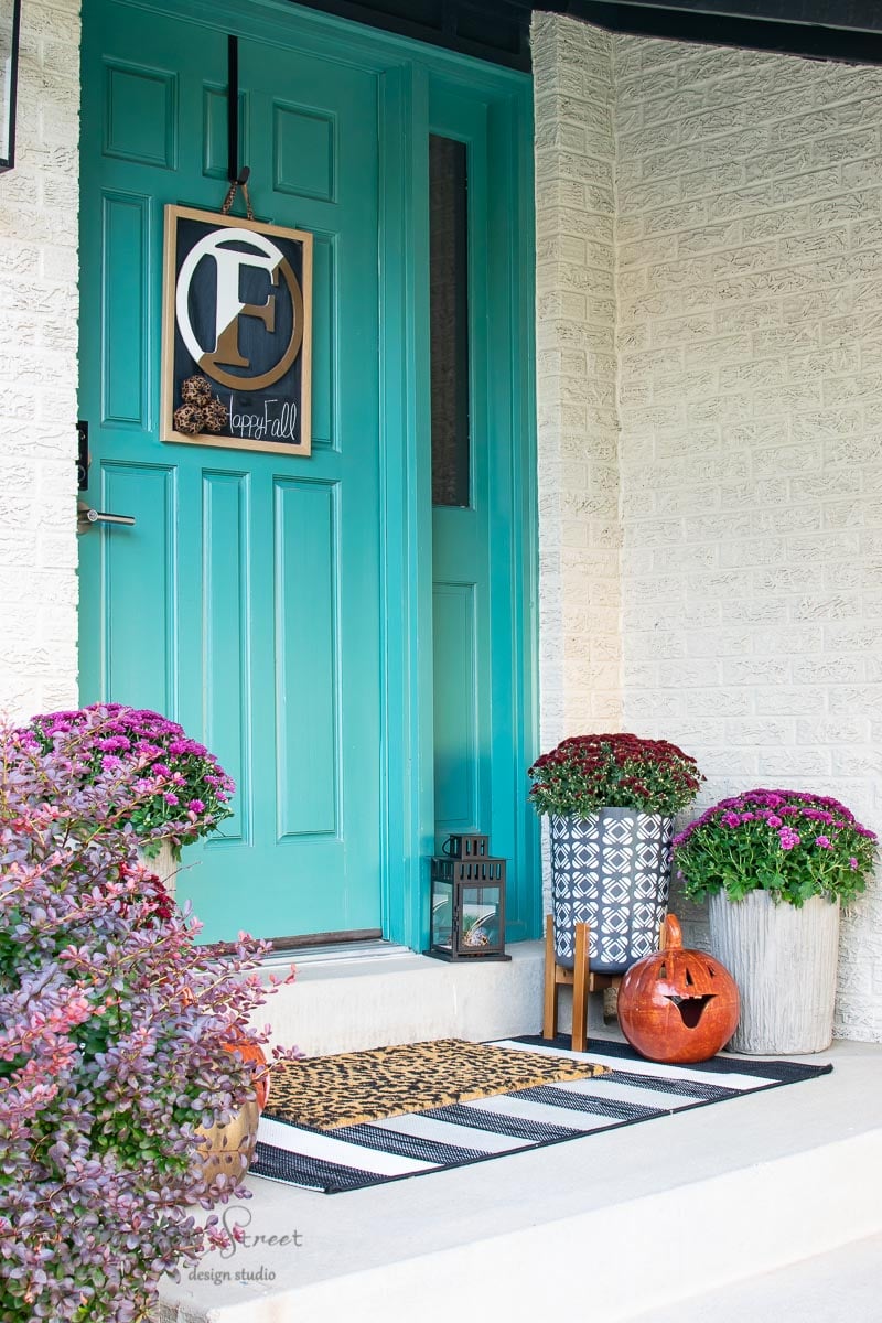 A turquoise front door with a framed "F" and "fall" sign hangs on it. The porch is adorned with a black lantern, potted purple flowers in a concrete planter, a decorative pumpkin, and a striped welcome mat.