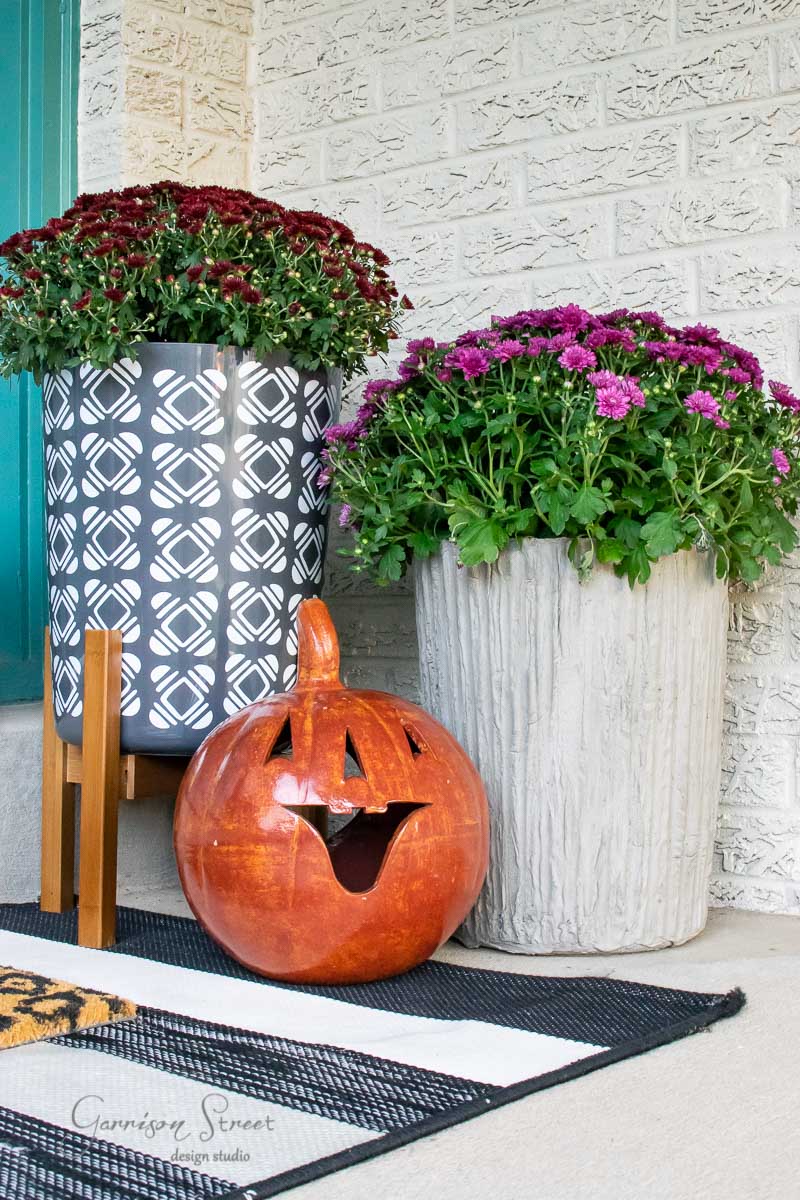 A porch decorated for fall showcases two large concrete planters brimming with red and purple flowers. Between them, a carved ceramic pumpkin with a smiling face adds charm. A black-and-white striped doormat is partially visible underneath.