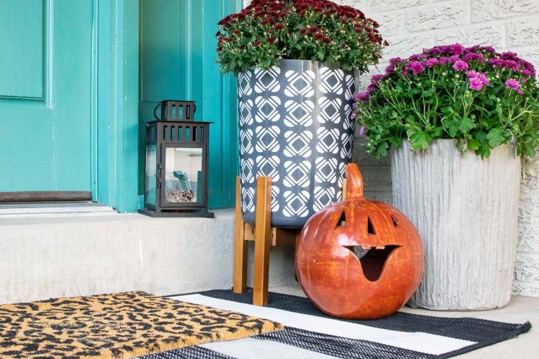 A porch adorned with red and purple flowers in concrete planters, complemented by a blue lantern and a ceramic pumpkin with a cutout face. The entrance features a turquoise door and a black-and-white checkered rug topped by a leopard print welcome mat.