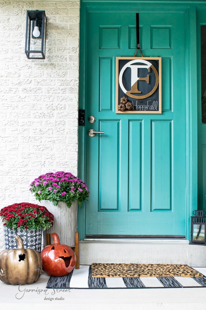 A turquoise front door decorated with a fall-themed wreath, featuring an "E" and pinecones. A concrete planter brimming with colorful flowers and decorative pumpkins lines the steps beside a striped doormat. To the left, a wall lantern casts a warm glow.