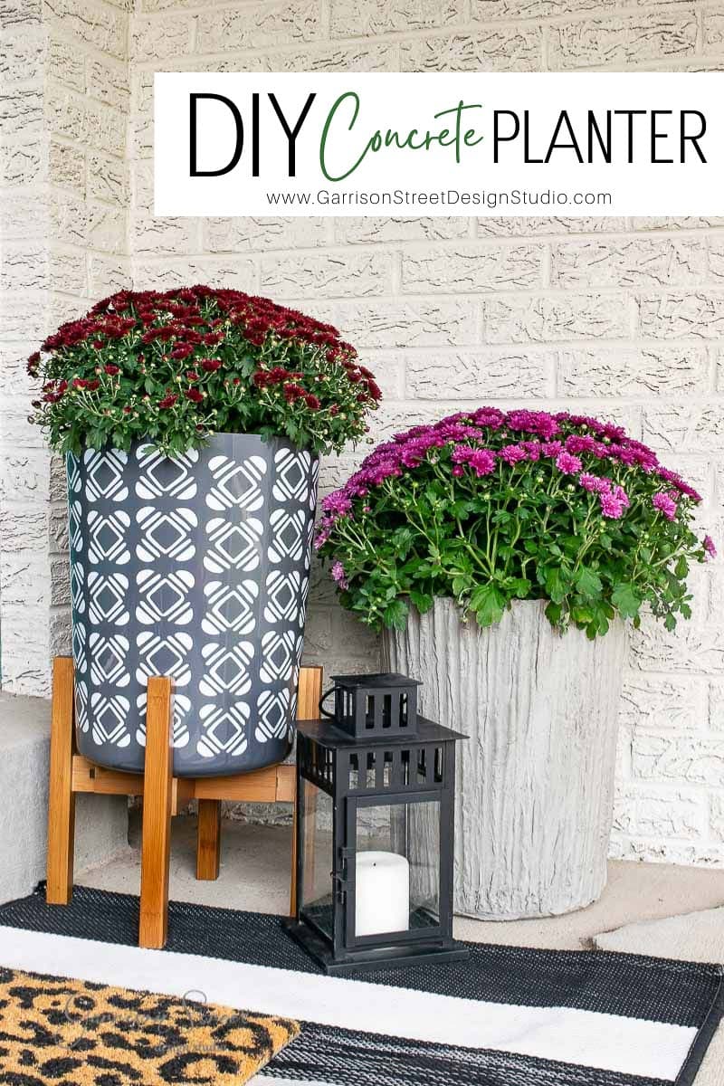 A porch with two large planters; one is dark gray with a geometric pattern on a wooden stand, and the other is a DIY concrete planter with rustic charm. Both are filled with vibrant, blooming flowers. A black lantern with a white candle sits in front.