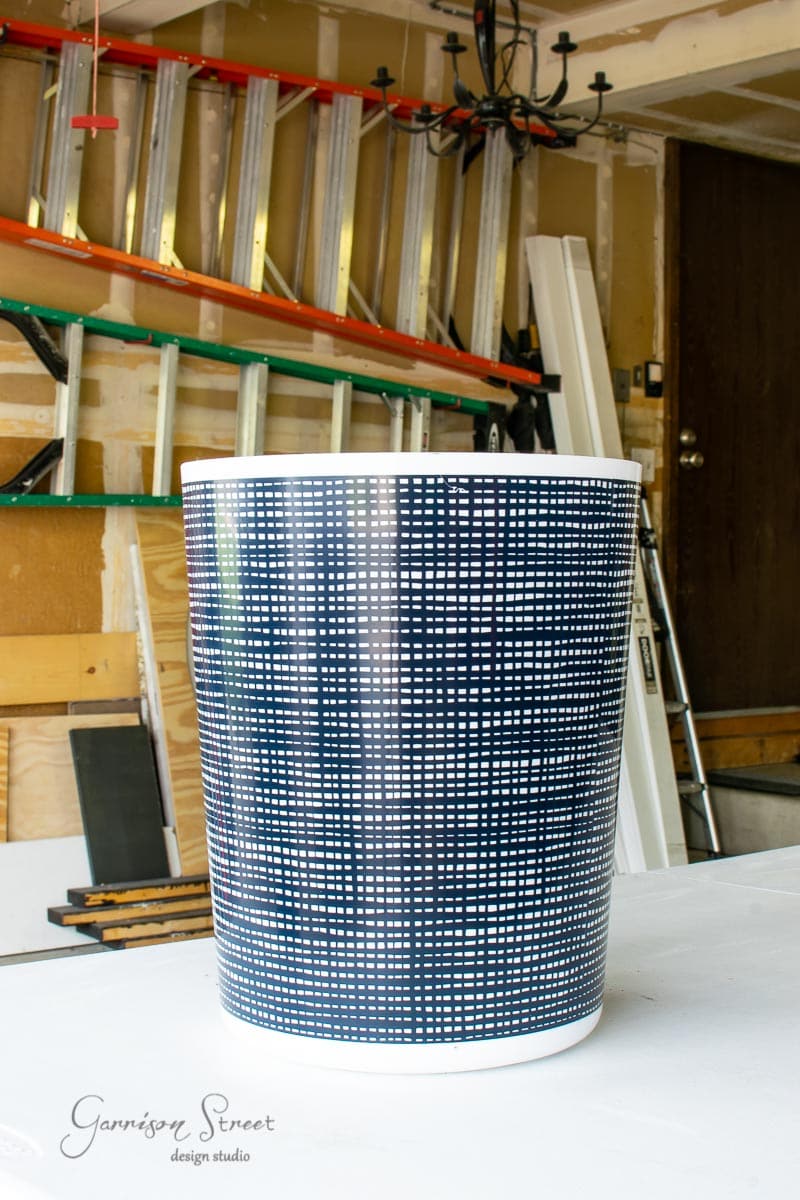 A black and white woven-patterned trash can sits on a white table next to a concrete planter in a garage. In the background, ladders are mounted on the wall, with various tools scattered around.
