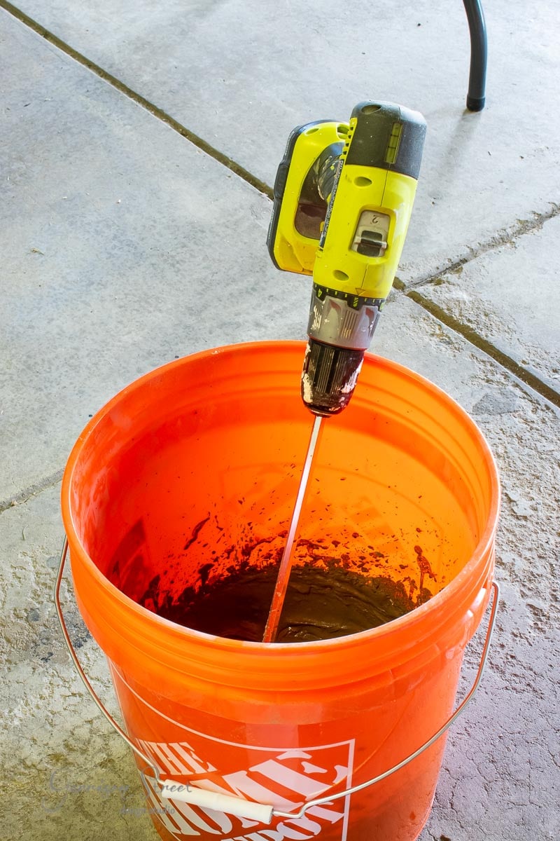 An orange bucket filled with a dark mixture, possibly for crafting a concrete planter. A power drill with a mixing attachment is poised in the bucket, indicating the concoction is being stirred. The scene unfolds on a concrete floor.