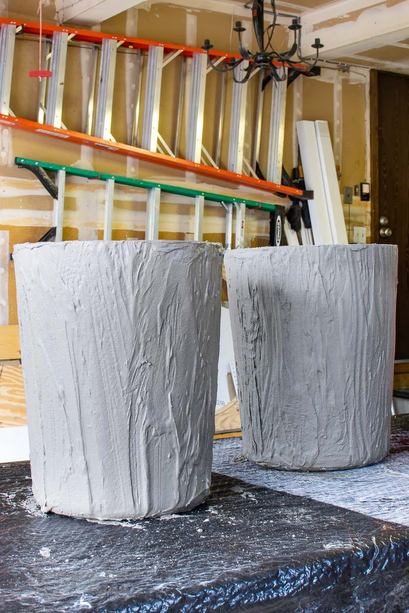 Two large concrete planters with a textured finish sit on a black plastic covering in a garage. In the background, various ladders and tools are mounted on the wall. A chandelier hangs from the ceiling, casting light on this industrial setup.