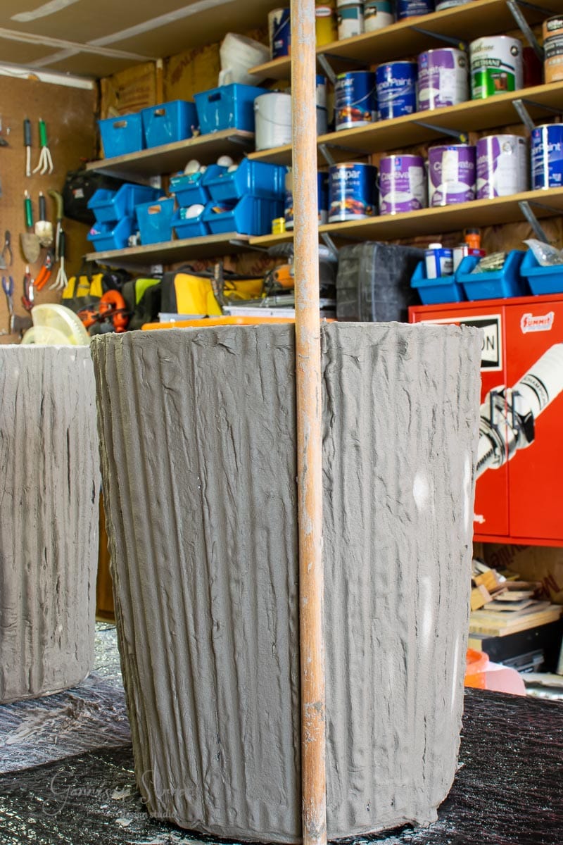 A large, textured cylindrical object resembling a concrete planter sits in the workshop, surrounded by shelves of paint cans, tools, and materials. A tall wooden pole stands beside it, hinting that this intriguing piece is still a work in progress.