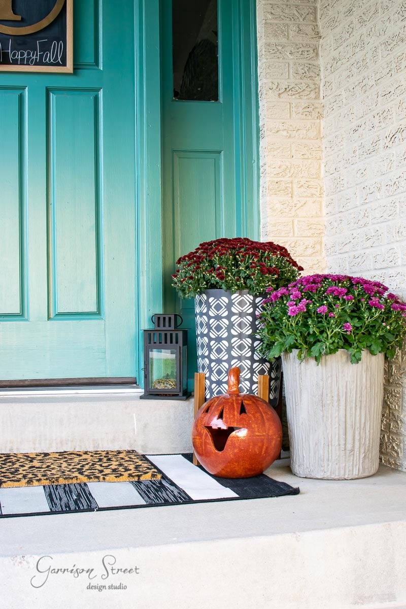 Front porch with a teal door, decorated for fall with a carved pumpkin, patterned lantern, and potted chrysanthemums in purple and red nestled in a concrete planter. A black and white doormat sits on the step, creating a festive autumn atmosphere.