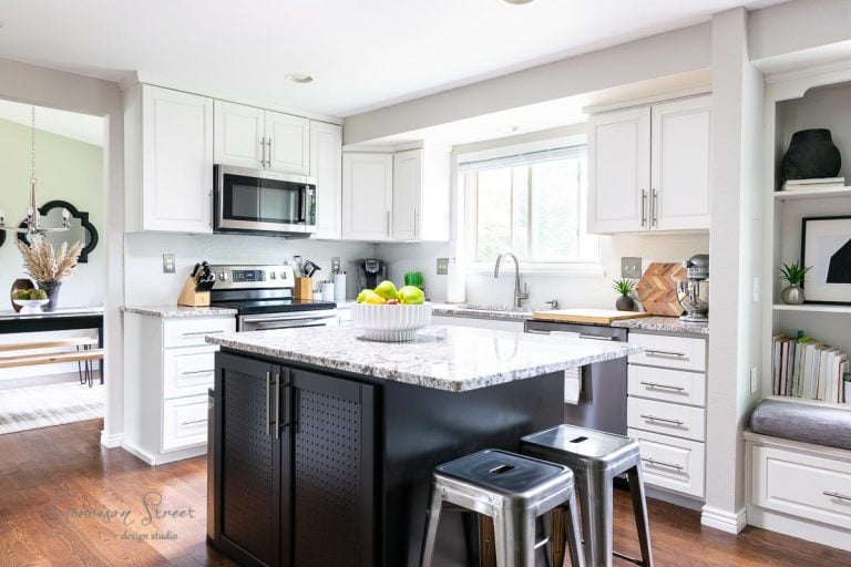 A modern kitchen showcases the power of paint with white cabinets, stainless steel appliances, and a central black island featuring granite countertops. Two metal stools are tucked under the island, and a bowl of green apples sits on top. Natural light streams in through a window.