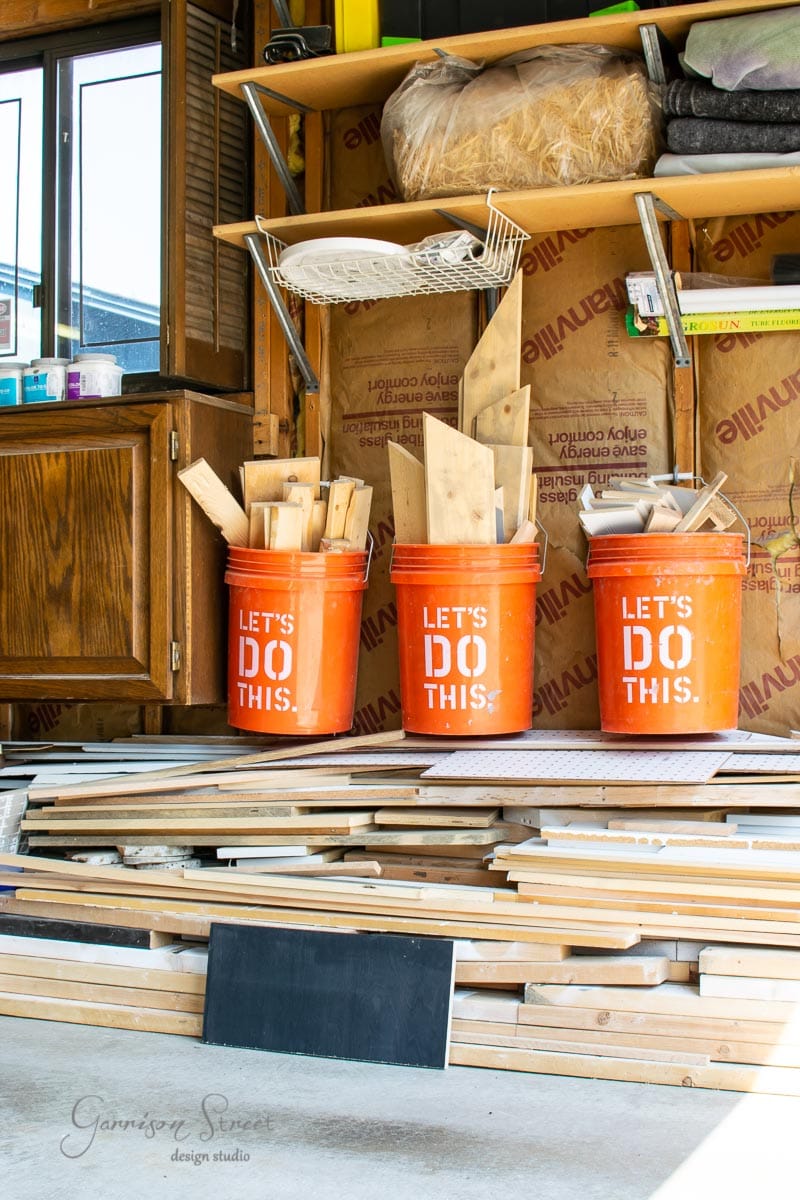 Three orange buckets labeled "LET'S DO THIS" are filled with wooden planks and scrap wood in a workshop. Behind them, insulating materials line the wall. A stack of various wood pieces is neatly arranged in front of the buckets.