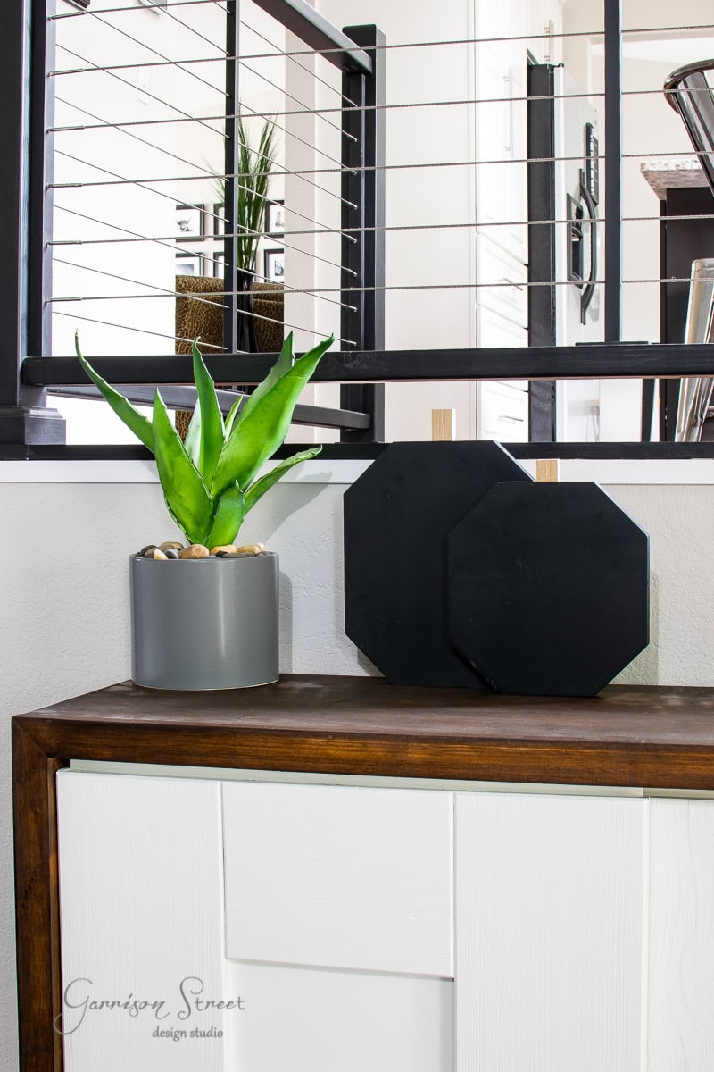 A modern interior scene showcases a sleek console table crafted from scrap wood, adorned with a small potted succulent and two black geometric decor pieces. In the background, a staircase with cable railing adds to the contemporary vibe.