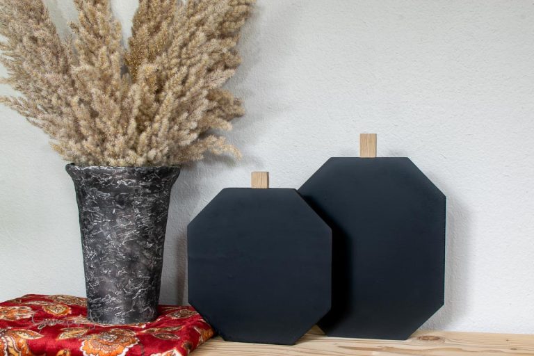 A decorative setup features a gray marbled vase holding tall dried grass, two black hexagonal cutting boards crafted from scrap wood with wooden handles in varying sizes, and a red floral cloth underneath, all elegantly placed on a wooden surface.
