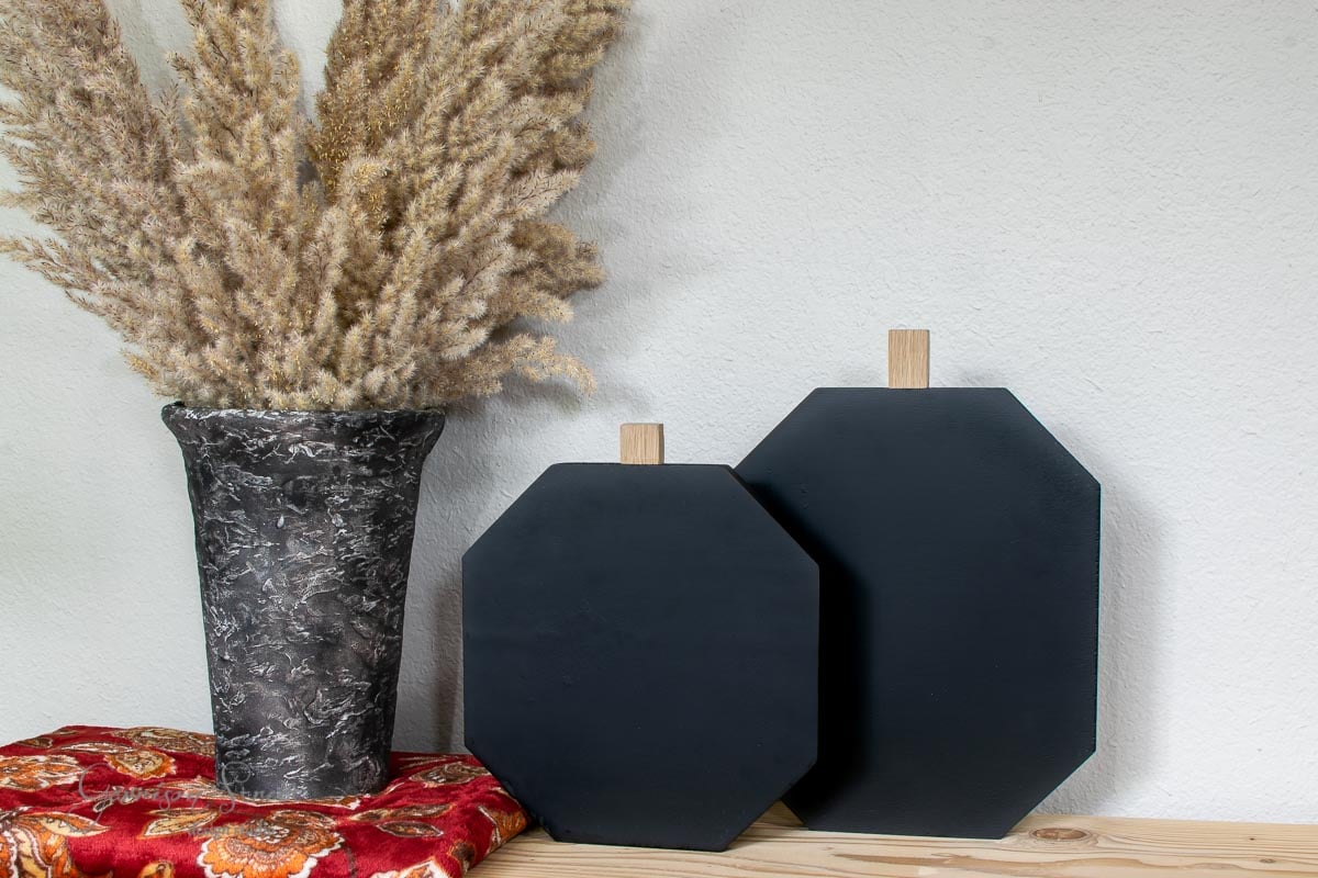A decorative setup features a gray marbled vase holding tall dried grass, two black hexagonal cutting boards crafted from scrap wood with wooden handles in varying sizes, and a red floral cloth underneath, all elegantly placed on a wooden surface.