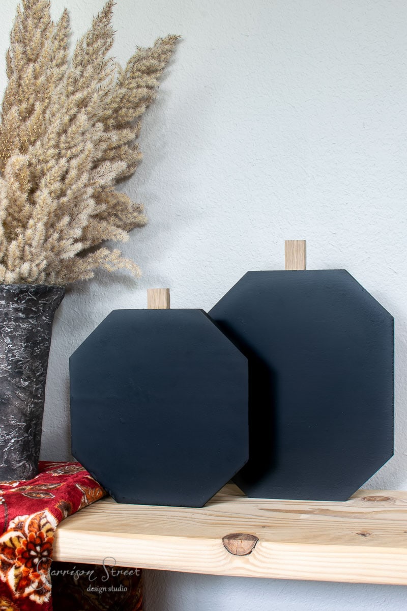 Two black, octagonal paddles crafted from scrap wood with wooden handles are displayed on a light wooden shelf. A vase with dried pampas grass and a red floral fabric rests beside them against a neutral wall.