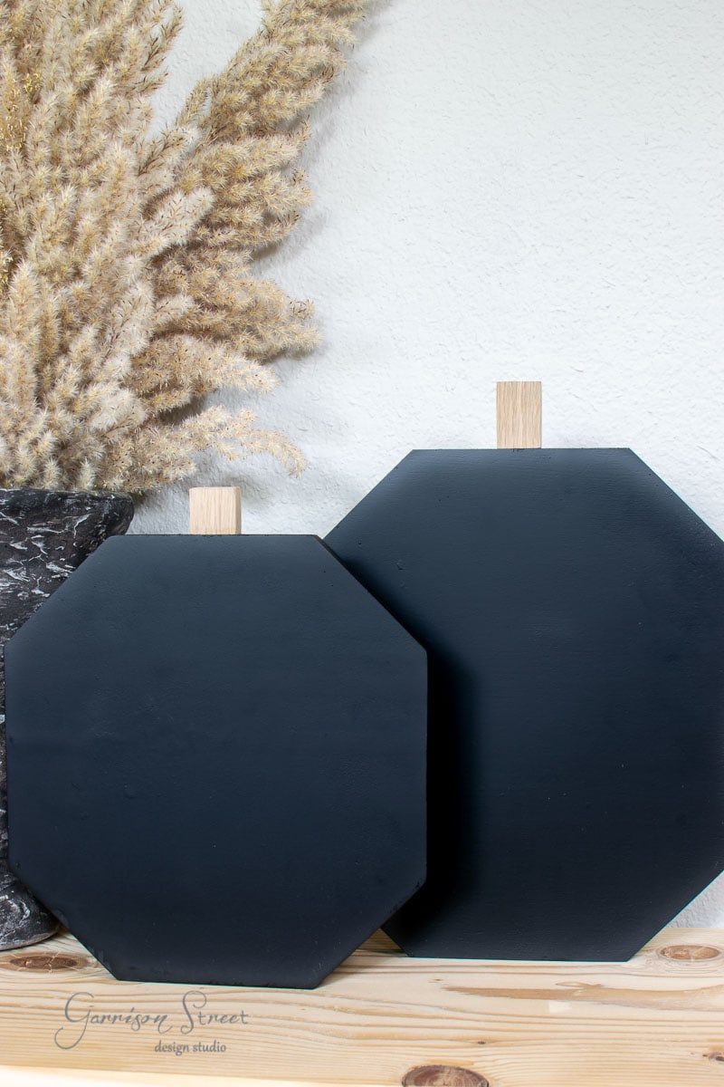 Two black, geometric-shaped cutting boards, crafted from scrap wood and featuring wooden handles, are propped against a light wall. They rest on a wooden surface next to a decorative vase filled with dried, feathery plants.