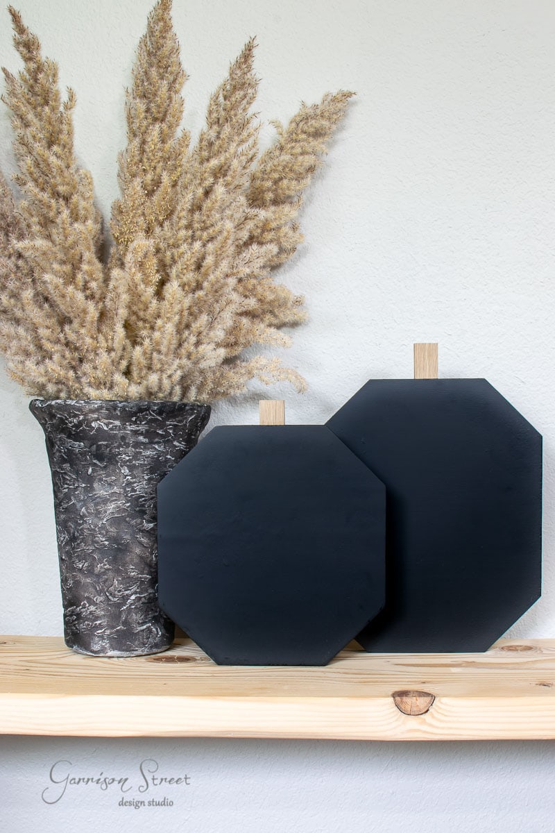 A decorative dried grass arrangement in a textured vase sits on a shelf crafted from scrap wood, next to two standing black chalkboard signs, one larger and one smaller. The brand name is subtly printed on the shelf.