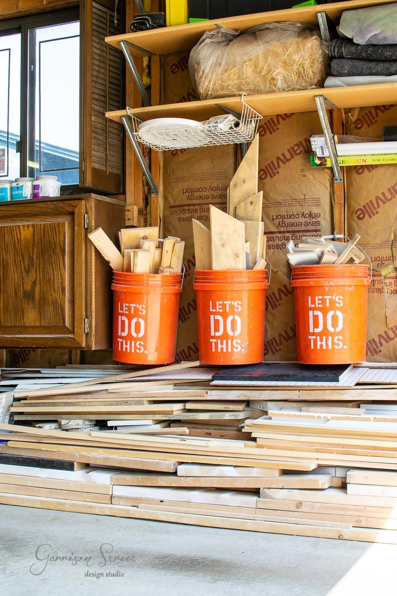 A garage workshop features three orange buckets labeled "Let's Do This," brimming with wooden planks and scrap wood. Stacks of lumber and various tools are neatly organized on a wooden shelf above. Insulation and wood cabinets add depth to the background.
