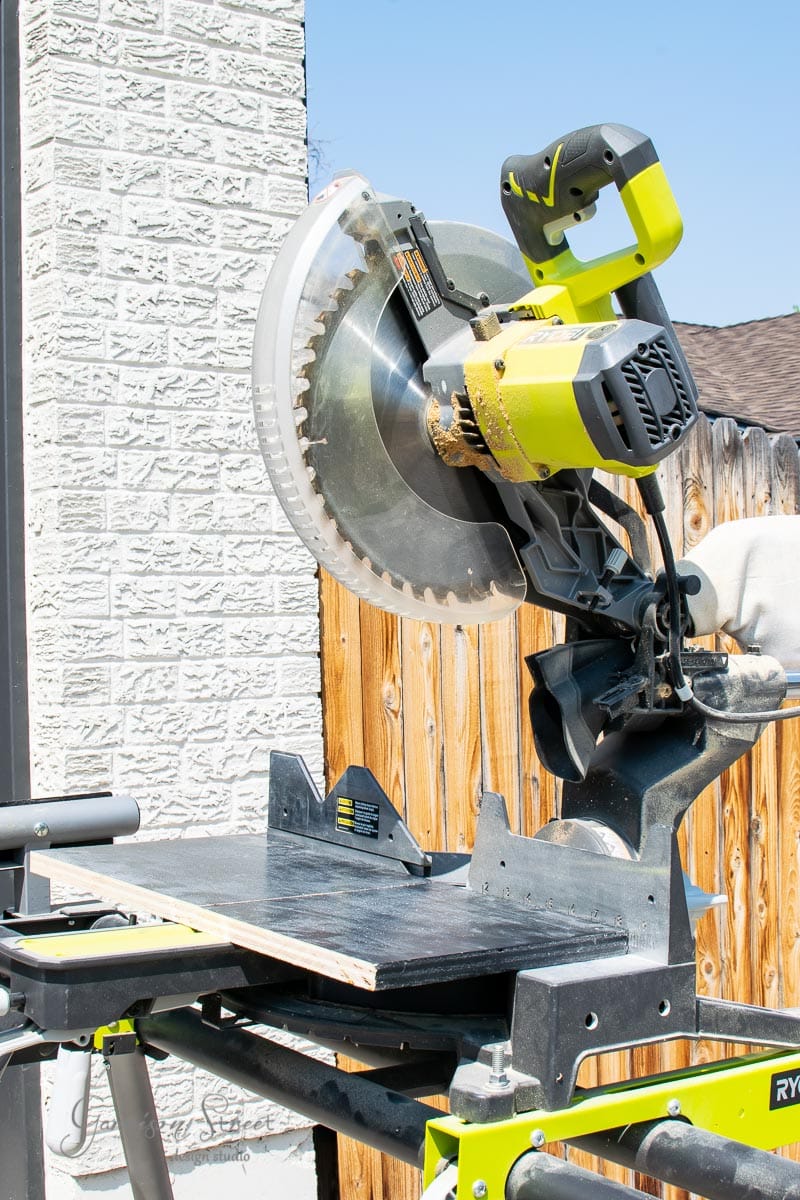 A miter saw with a bright green and black design is mounted on a workbench surrounded by scrap wood. It sits next to a brick wall and wooden fence, enjoying the backdrop of a clear blue sky.