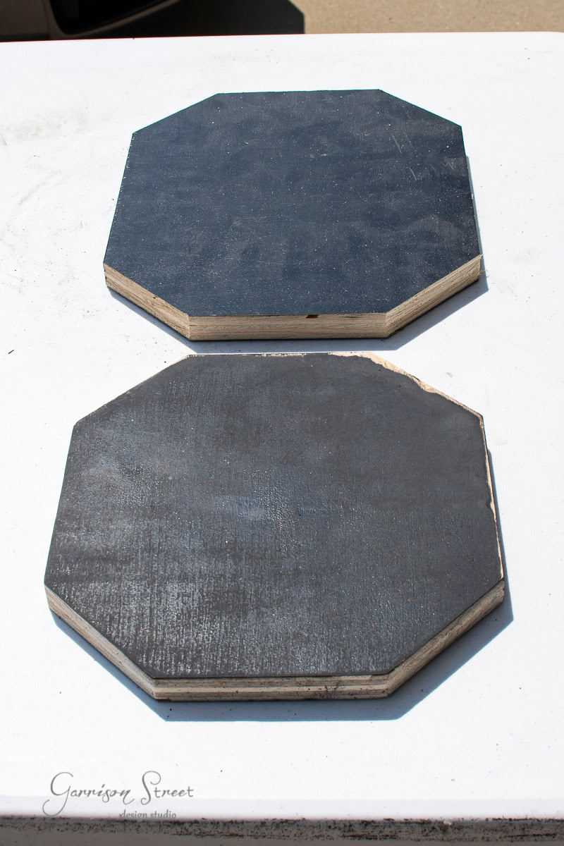 Two octagonal boards made from scrap wood are placed on a white surface. Painted black, they show a slightly weathered look with visible grain texture. The top board appears slightly smaller than the bottom one.