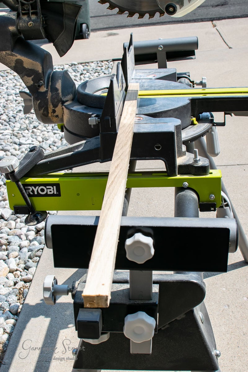 A wooden plank, perhaps salvaged from scrap wood, is positioned on a Ryobi miter saw stand, ready for cutting. The stand is set up on a paved area with decorative stones nearby. One corner of the miter saw blade can be seen in the background.