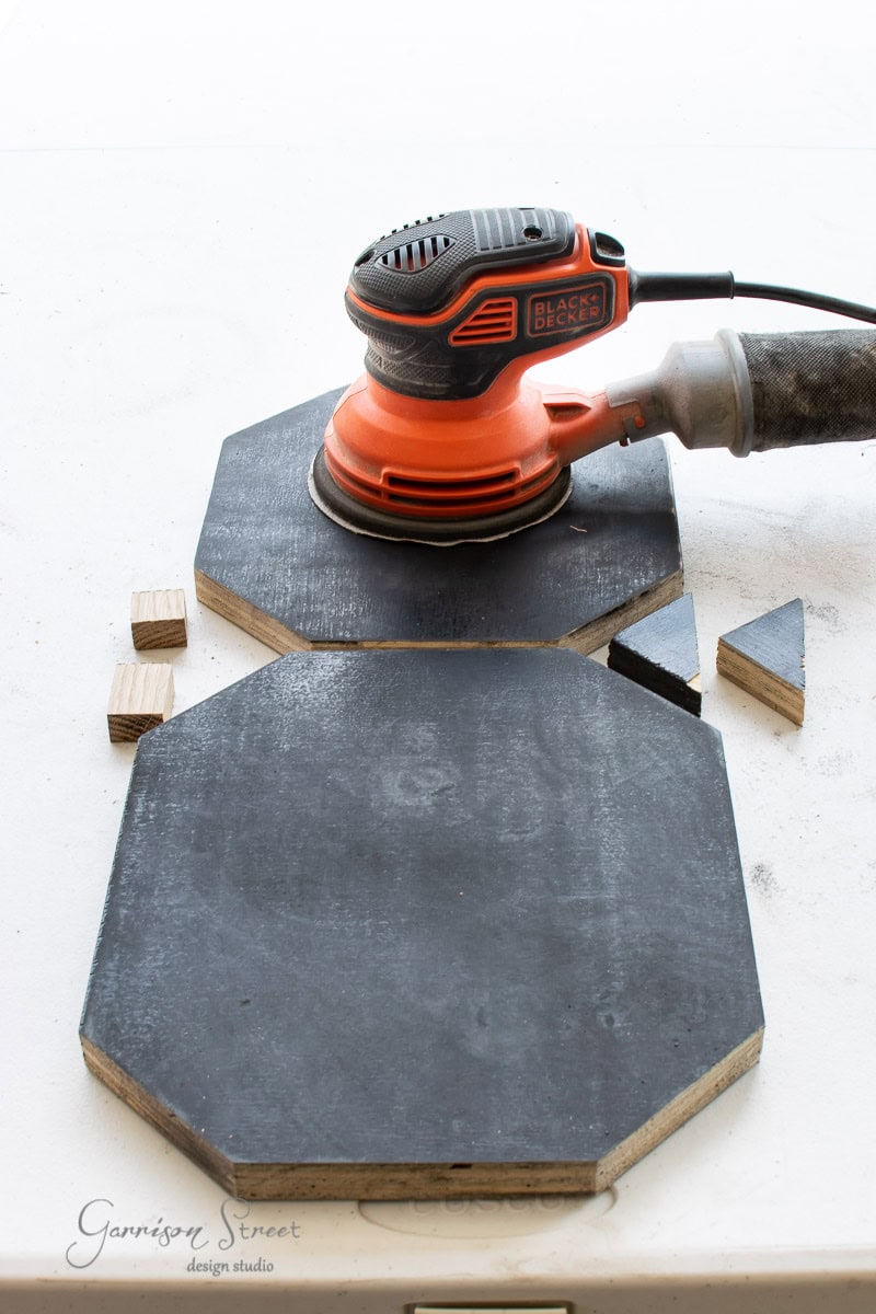 An orange and black electric sander rests on a black wooden hexagonal surface. Nearby, small wooden blocks and triangular pieces of scrap wood are scattered. The background is a white surface.