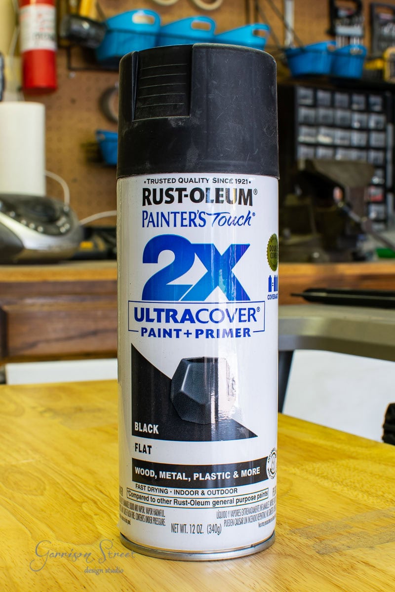 A can of Rust-Oleum Painter's Touch 2X Ultra Cover Paint + Primer in black sits on a workshop's wooden surface, surrounded by scrap wood, tools, and shelves in the background.