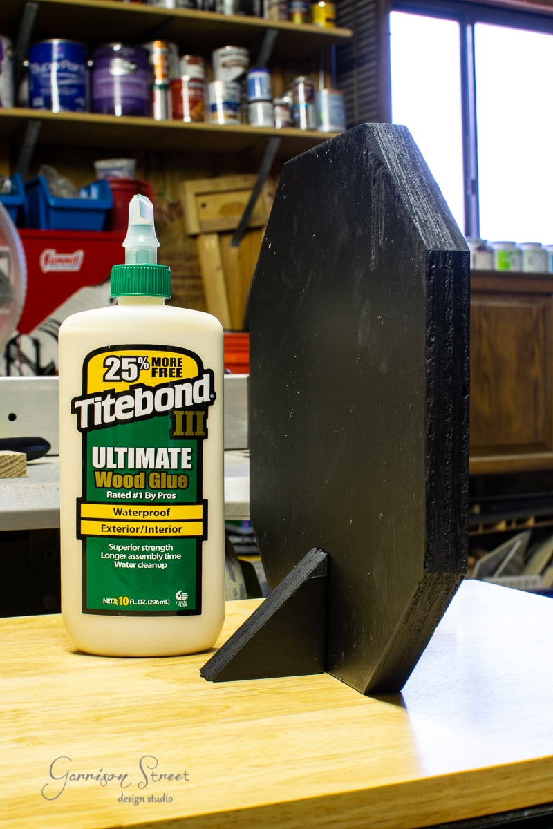 A bottle of Titebond III Ultimate Wood Glue sits on a workbench beside a black painted wooden piece, and scrap wood is scattered around. In the background, shelves brimming with various tools and materials create a bustling workshop setting.