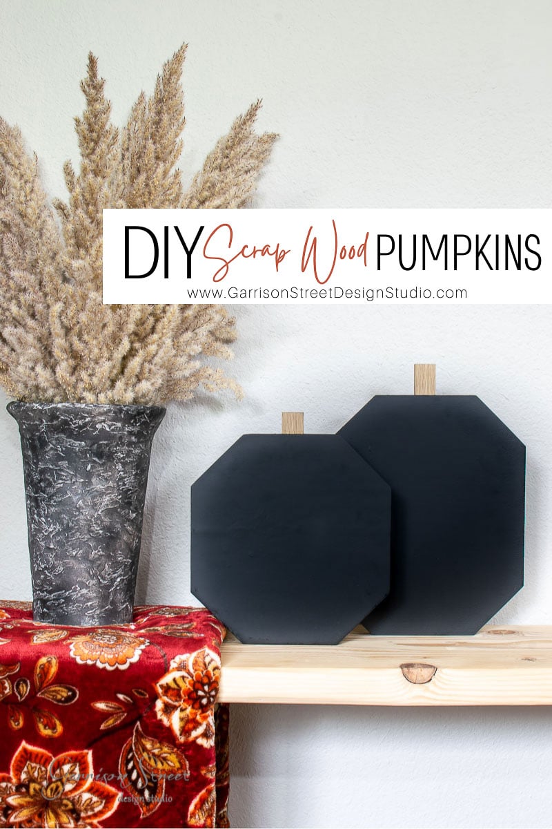 A decorative setup showcases two black octagonal wooden pumpkins crafted from scrap wood on a shelf, beside a tall vase of pampas grass. A red patterned cloth drapes elegantly over the shelf. Text reads "DIY Scrap Wood Pumpkins" with a website link below.