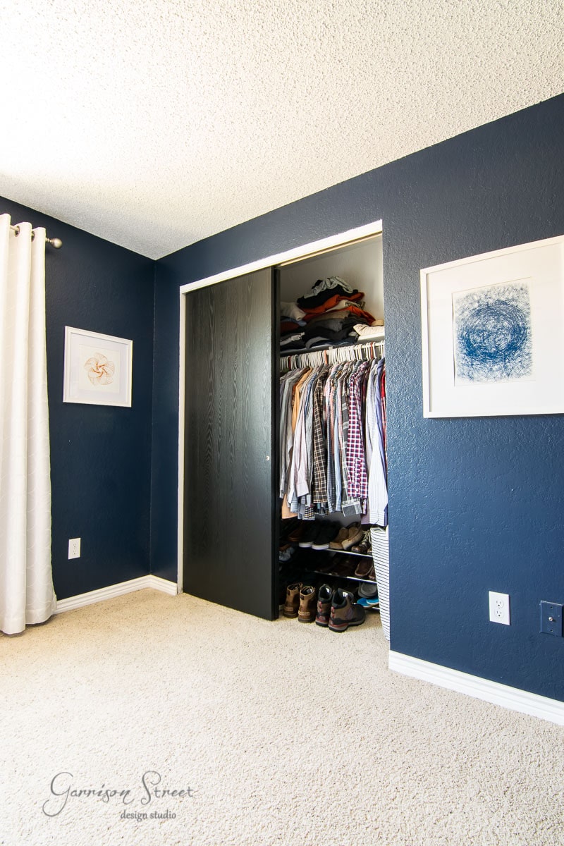 A neatly organized closet with shirts and jackets hanging on a rod, shoes lined up at the bottom, exudes a modern boho touch. The dark sliding doors complement the dark blue walls, white curtains, and framed art in this stylish space.