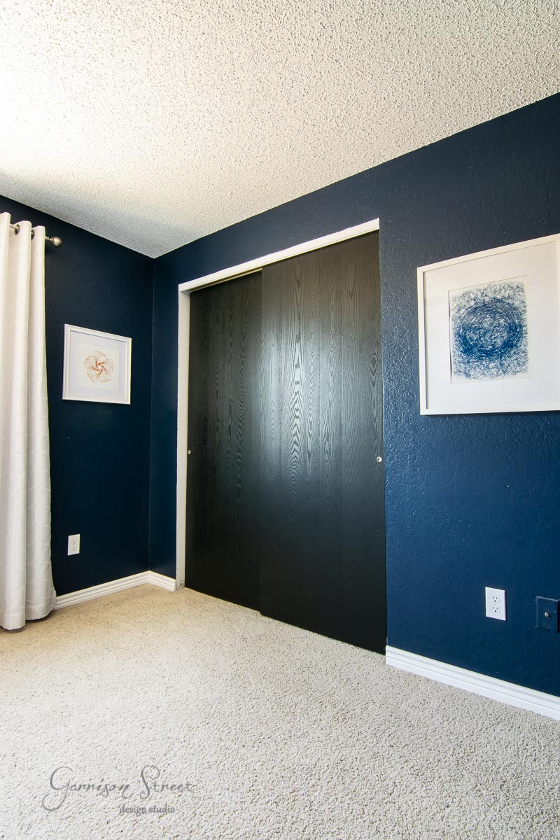 The modern boho room features dark blue walls and a white carpet, exuding a chic vibe. Framed art adorns the walls above dark-colored sliding closet doors, while white curtains softly drape from one side. It's both stylish and inviting, perfect for either a cozy retreat or an artsy bunk room.