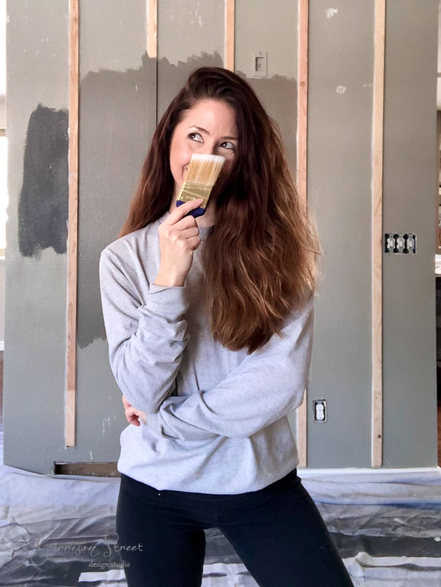 A woman with long hair in a gray sweatshirt stands in front of an unfinished wall, holding a paintbrush near her face. The wooden studs and patches of gray paint evoke a modern boho vibe, suggesting a creative DIY activity or the transformation of a future bunk room.