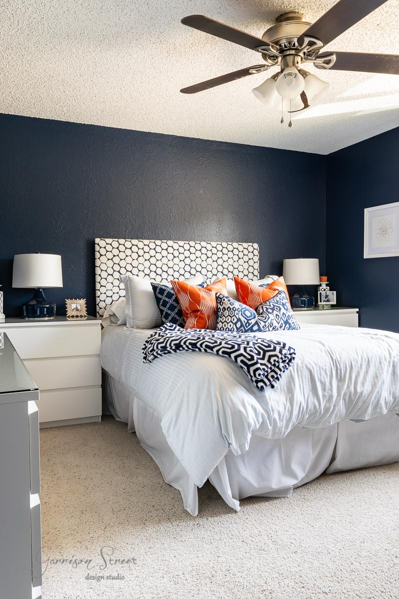 A bunk room with a dark blue accent wall features a bed with a patterned gray and white headboard. The bed is dressed with white linens and decorative pillows in orange and navy boho patterns. A ceiling fan is above, and two nightstands with lamps flank the bed.