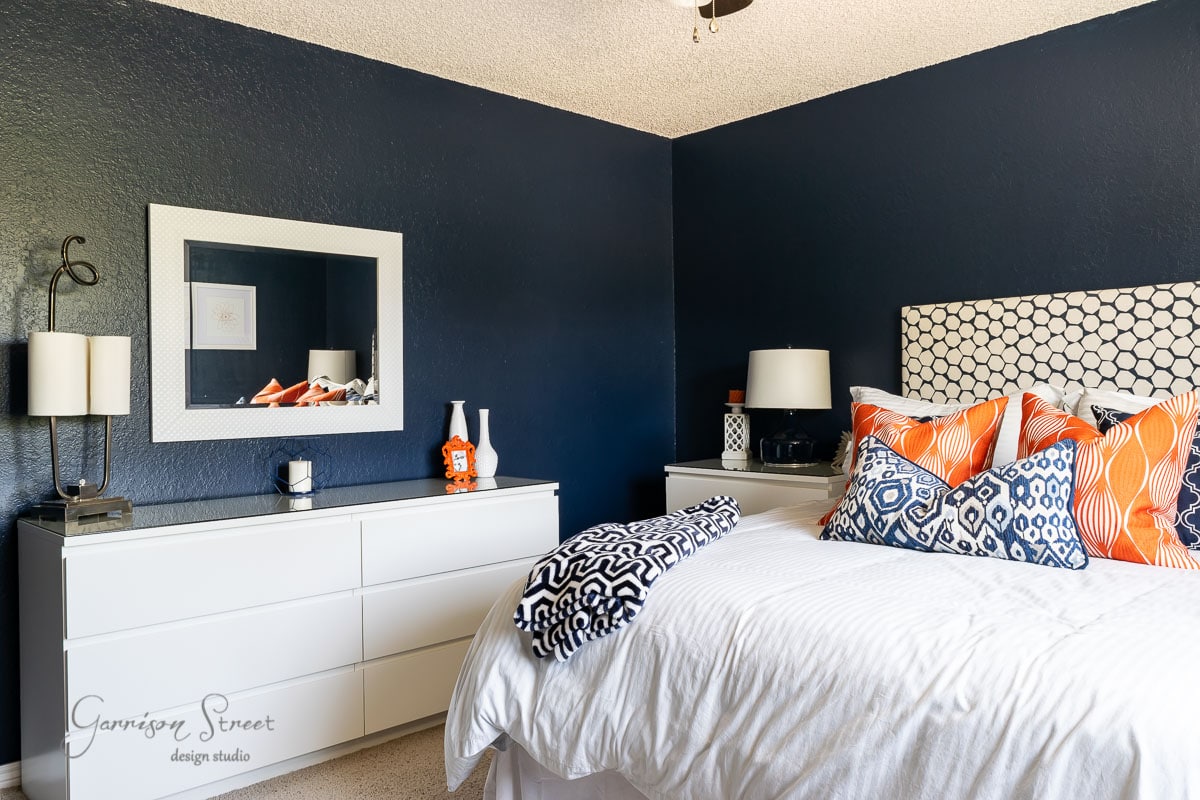 A bedroom with dark blue walls embodies a modern boho vibe, featuring a white bed adorned with patterned pillows in orange, white, and blue. A white dresser with a mirror sits to the left, decorated with lamps and a vase. The room exudes a cozy yet chic ambiance.