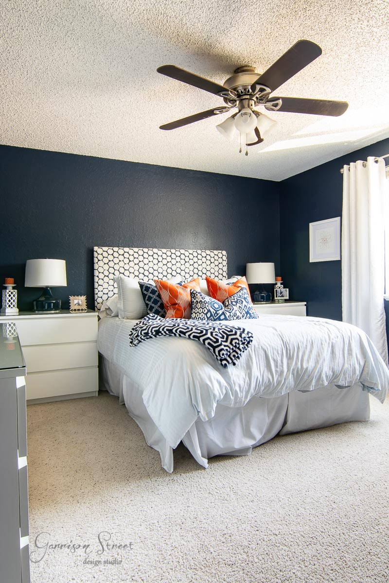 The bedroom boasts a modern boho vibe with a white bed, patterned pillows, and a geometric headboard. Navy blue walls contrast beautifully with the beige carpet. Two white nightstands hold lamps, while a ceiling fan with lights hangs above. Sunlight streams through the window's white curtains.