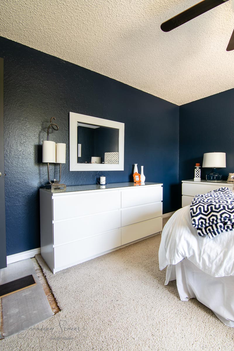 Modern boho bedroom with dark blue walls and a textured white ceiling. A large white dresser holds two ceramic vases and a lamp, with a rectangular mirror mounted above. Nearby, a bed with white bedding and a patterned navy throw completes the stylish ensemble.