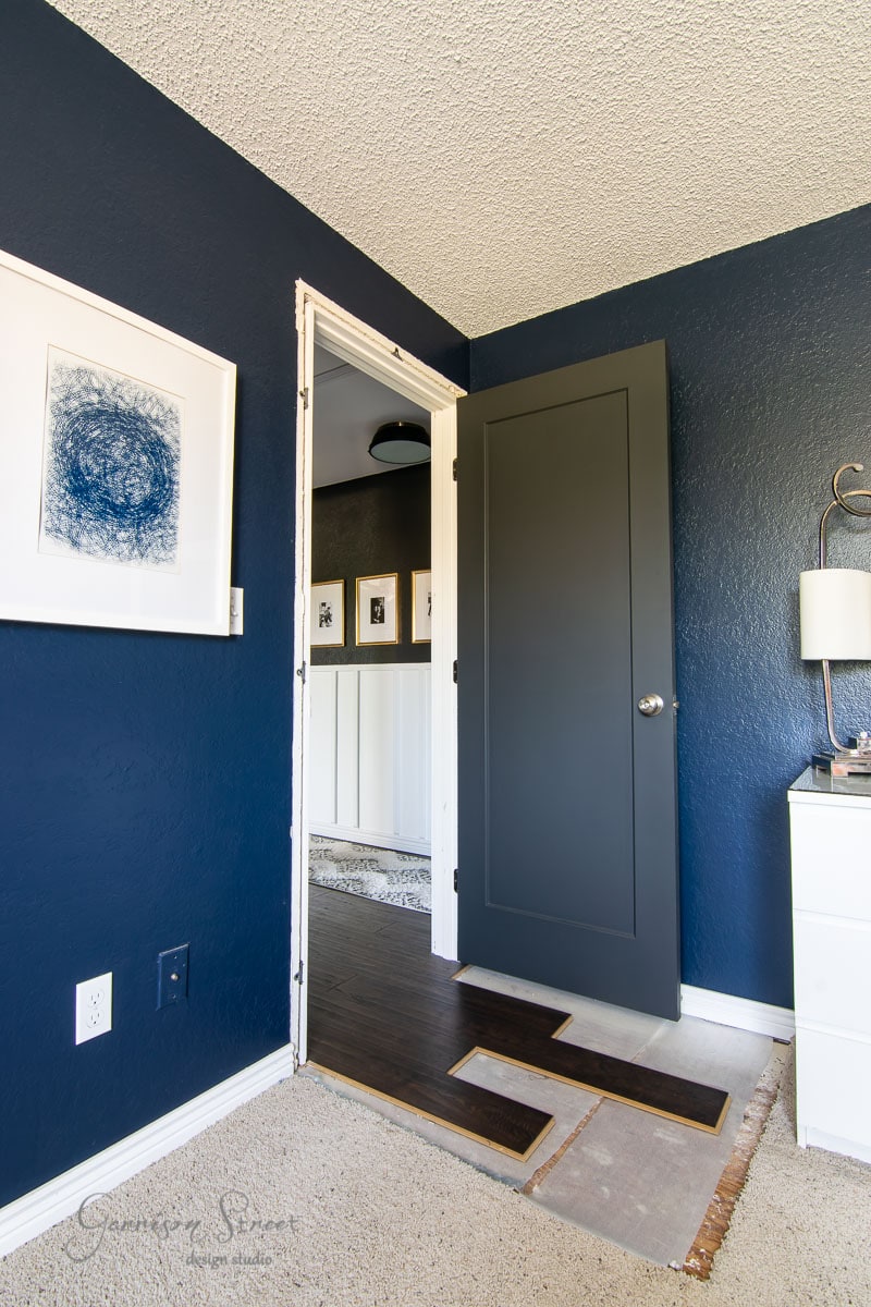 A modern boho room with dark blue walls and a partially open dark gray door. The walls showcase framed abstract artwork. The floor blends carpet and laminate panels. A white dresser with a lamp is visible to the right, adding charm to this eclectic space.