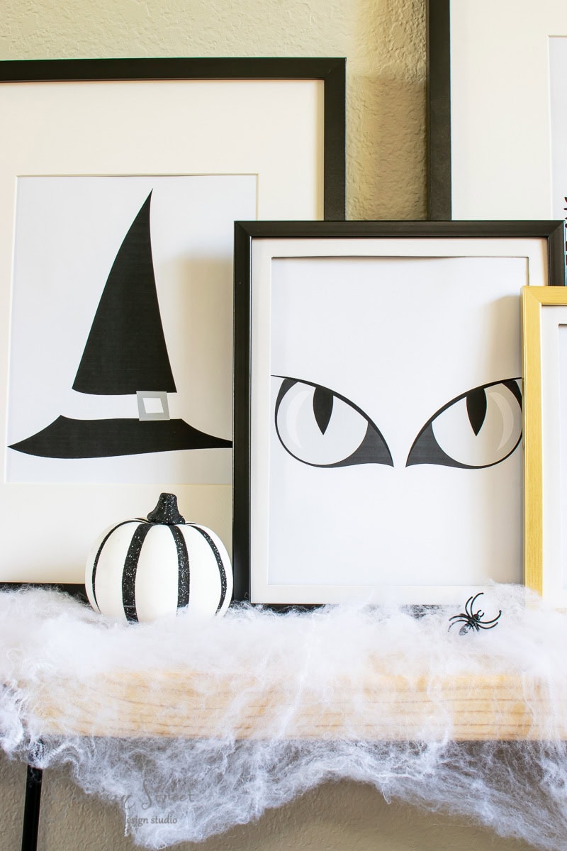 A Halloween-themed display features framed art of a witch's hat and black cat eyes. A small black and white pumpkin sits on a shelf adorned with faux spiderwebs and a plastic spider.