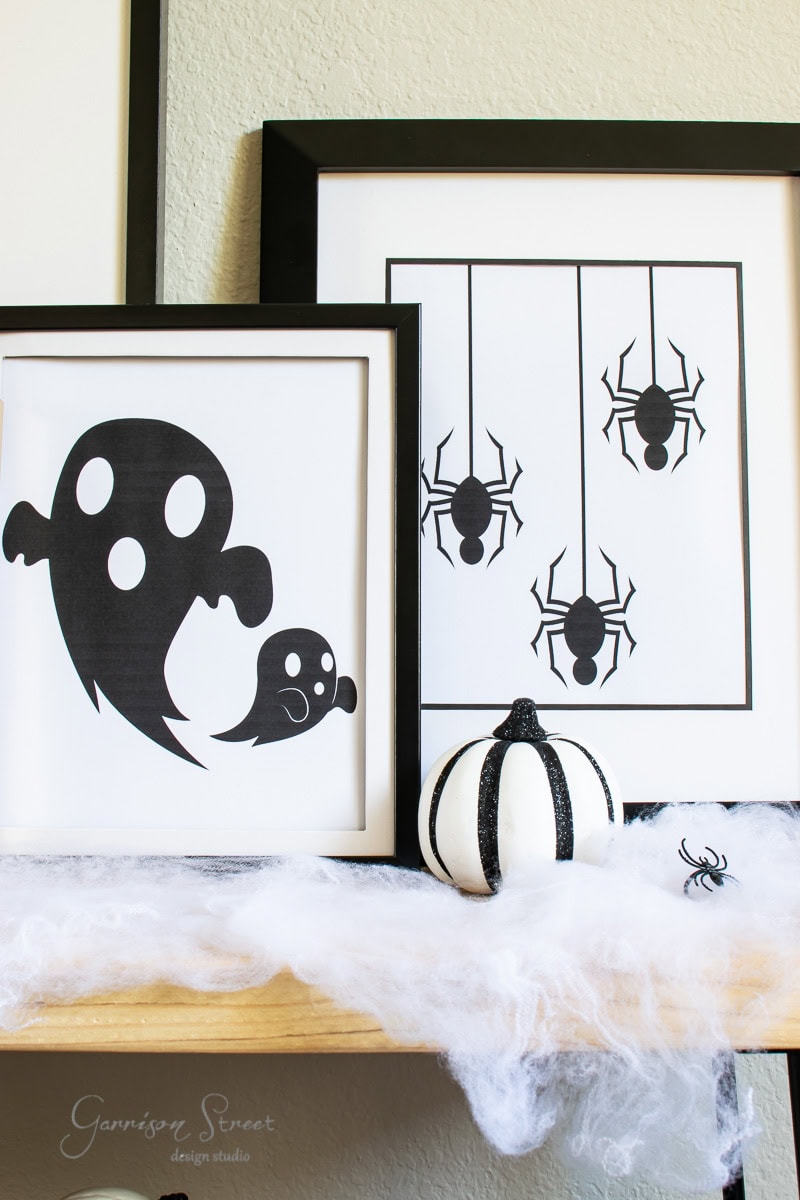 Halloween-themed decor featuring framed black and white prints of a ghost, hanging spiders. A small white pumpkin with black accents sits on a cobweb-covered surface, accompanied by a tiny decorative spider.