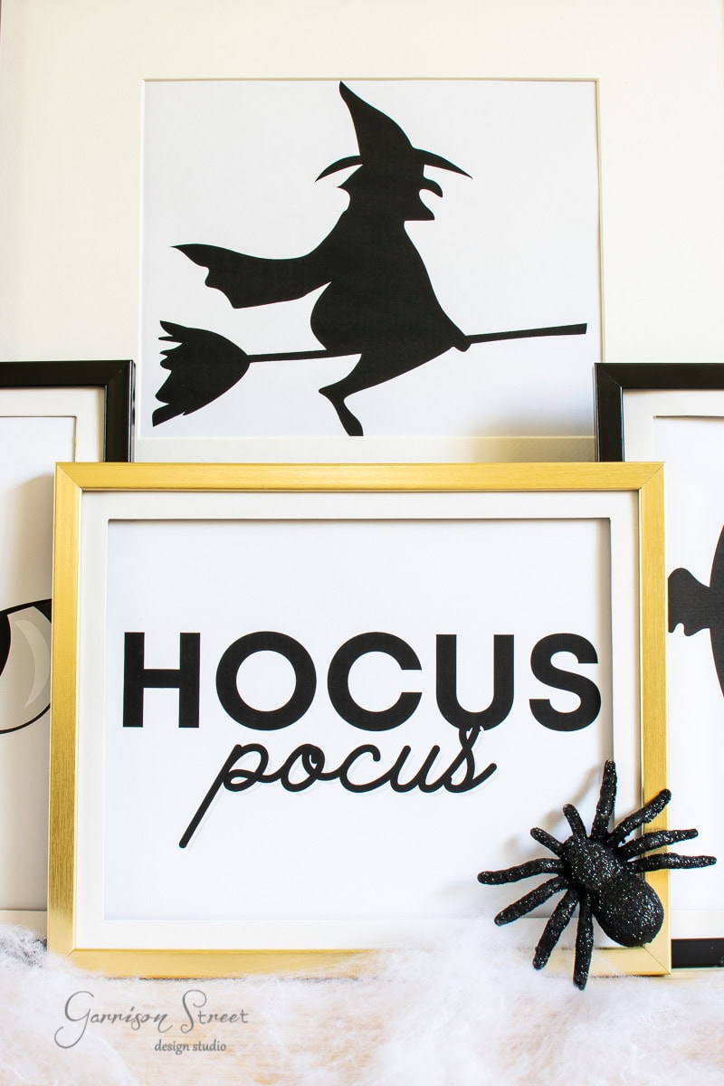 A framed print features the words "Hocus Pocus" alongside a black glittery spider decoration in the bottom corner. Behind it, another frame displays a silhouette of a witch on a broomstick, both set against a soft, white background.