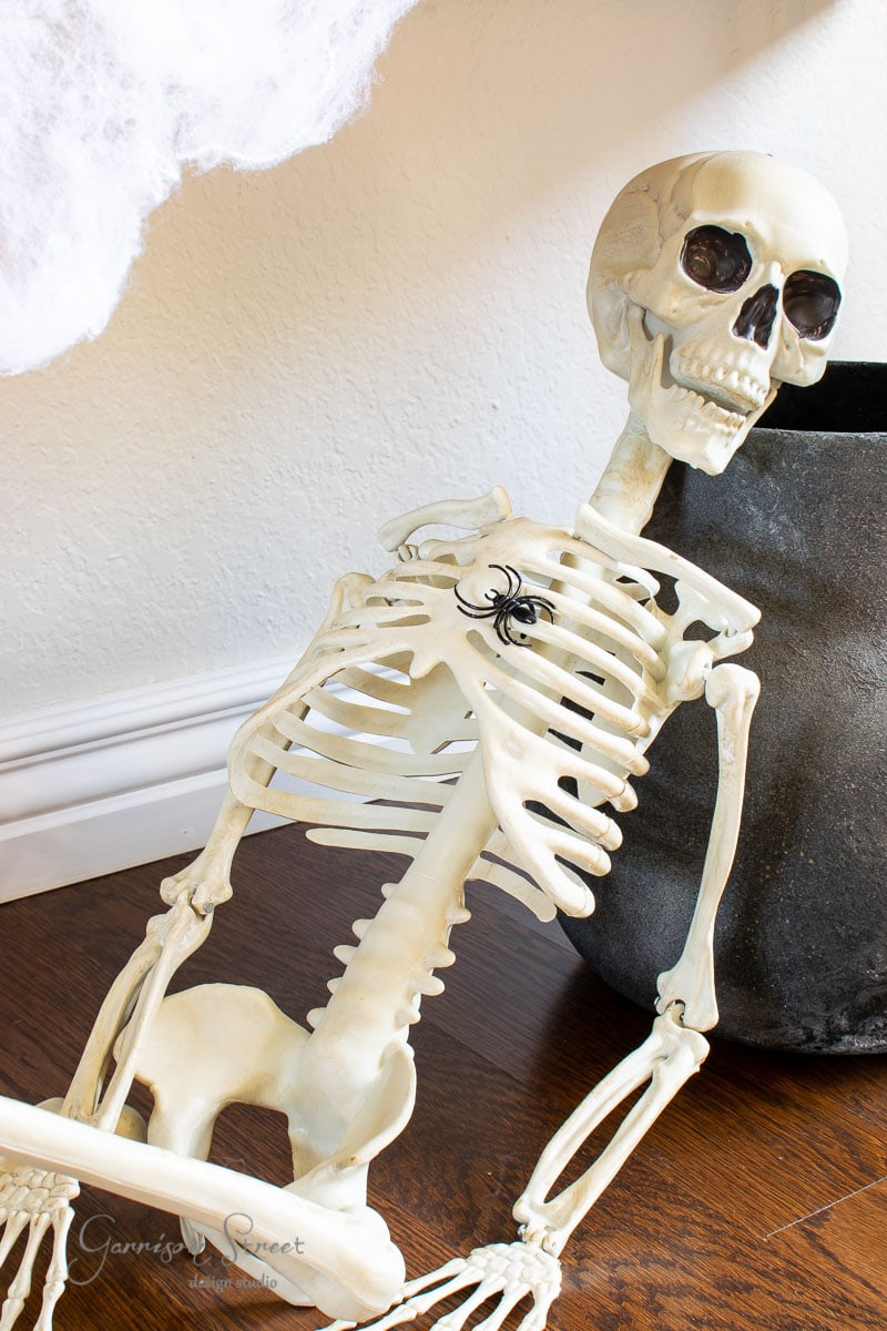 A toy skeleton leans against a dark pot on a wooden floor, with a small black spider perched on its ribcage. White fabric drapes partially visible in the upper left corner, completing the eerie scene.