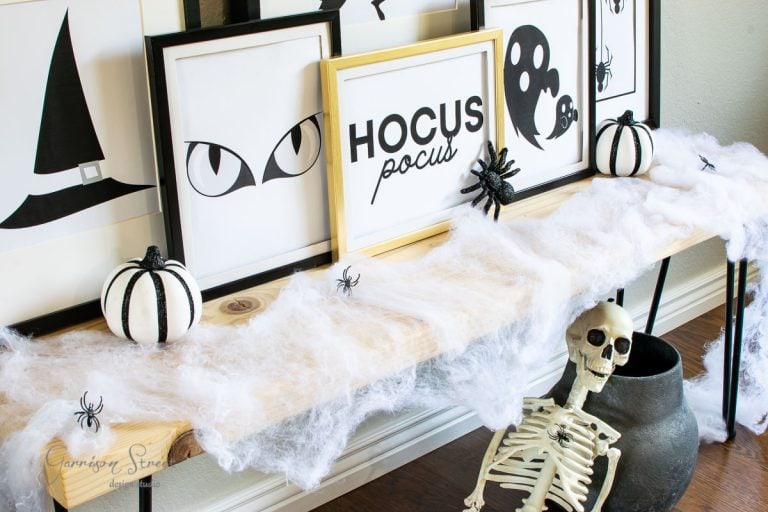A Halloween-themed display on a table features framed art, including witch eyes and "Hocus Pocus" text. Decorative black and white pumpkins, spiders, and fake cobwebs adorn the table. A skeleton sits beside a black pot on the floor, surrounded by charming Halloween sign printables.