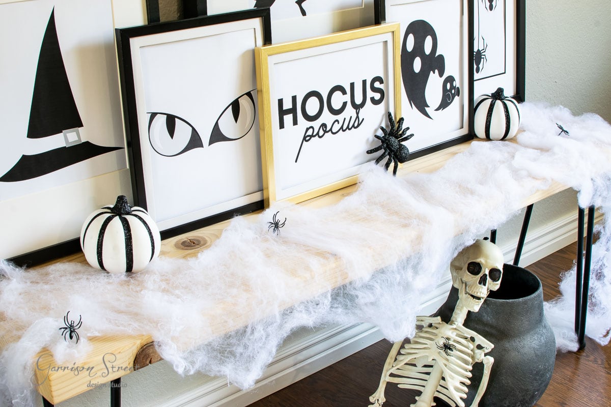 A Halloween-themed display on a table features framed art, including witch eyes and "Hocus Pocus" text. Decorative black and white pumpkins, spiders, and fake cobwebs adorn the table. A skeleton sits beside a black pot on the floor, surrounded by charming Halloween sign printables.