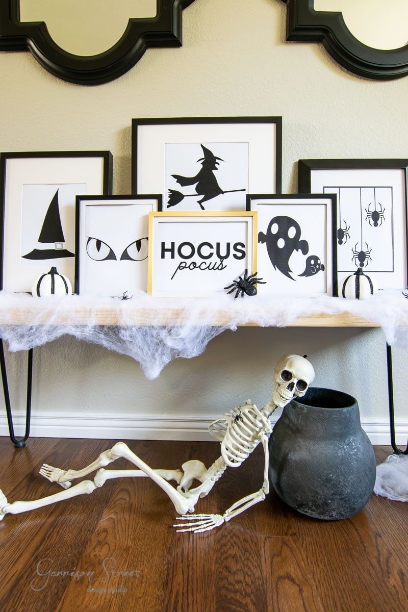 A Halloween-themed room features framed silhouettes of a witch, hat, cat eyes, and spiders. A "Hocus Pocus" sign adds charm. A skeleton reclines by a large black cauldron amid cobwebs on the table.