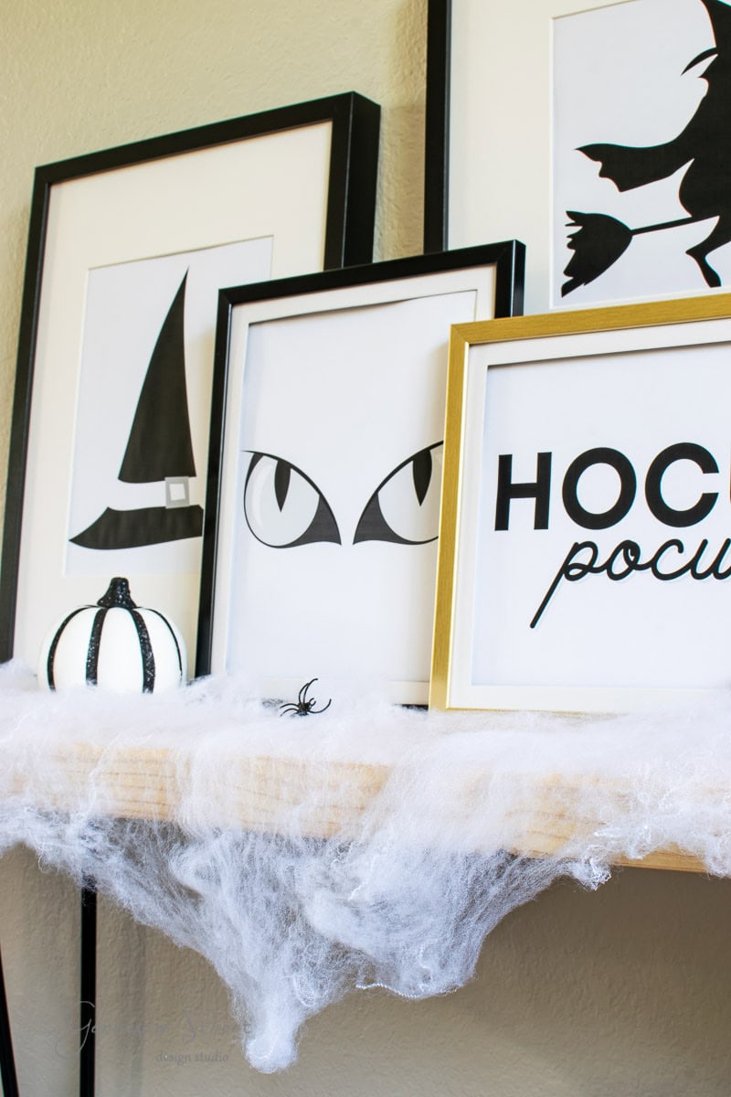 A shelf decorated for Halloween features framed prints, including a witch's hat, cat eyes, and a silhouette of a witch on a broom. A black and white pumpkin and faux cobwebs with a small spider enhance the spooky theme.