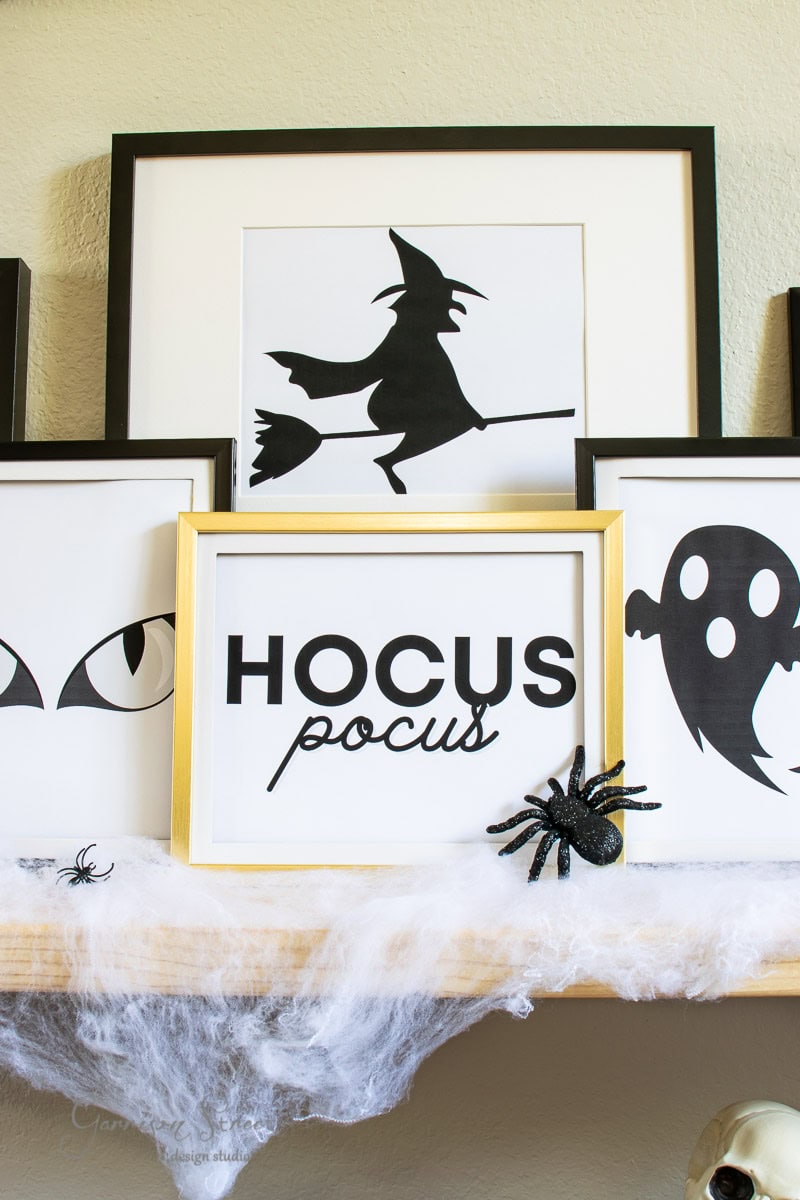 A shelf showcases framed Halloween-themed prints, featuring a witch on a broomstick, a ghost, and the text "HOCUS pocus." Faux cobwebs and plastic spiders enhance the spooky display.